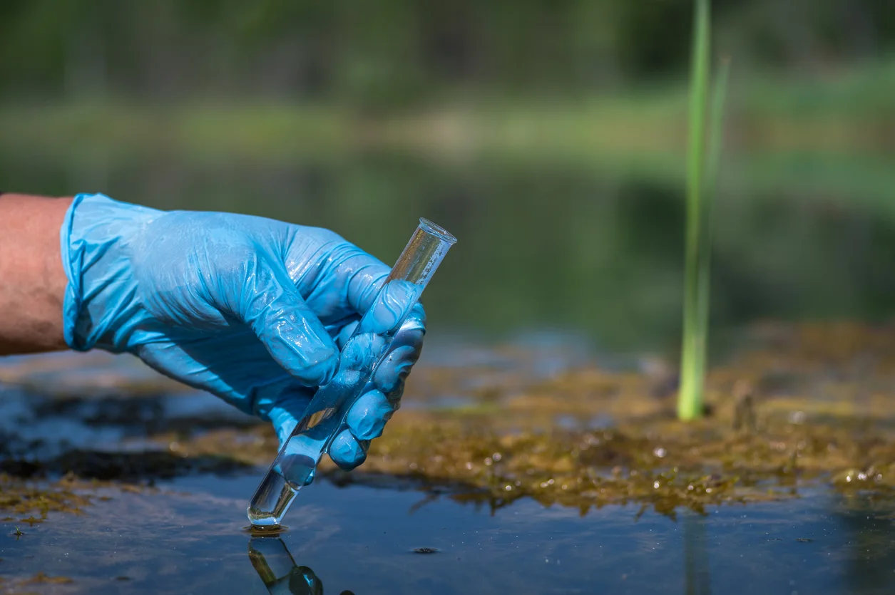 An image of a person's hands wearing blue safety gloves holding a test tube to test water from a pond or lake representing PFAS contamination and toxic exposure claims litigated by Trident Law HQ