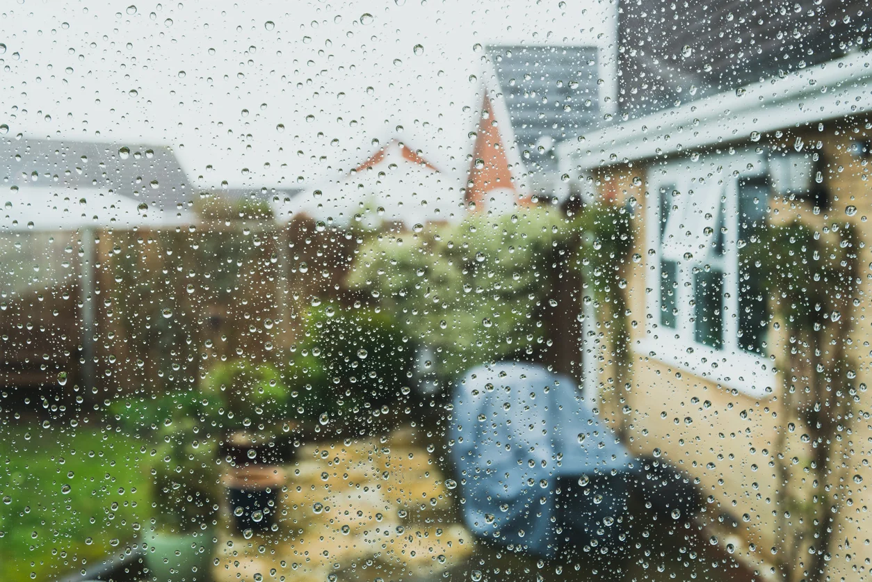 An image of a window covered raindrops showing the view of a yard with houses representing widespread storm destruction and storm damage claims handled by Trident Law HQ