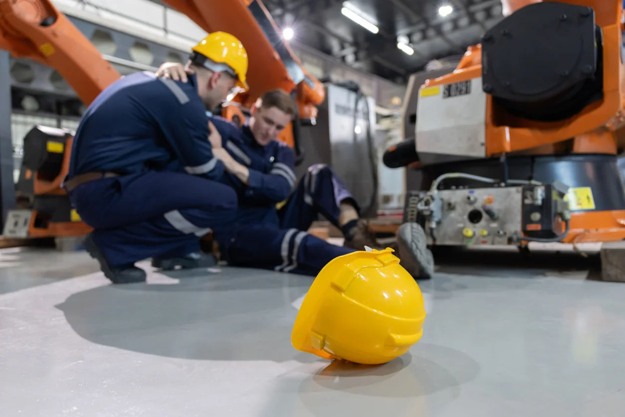 A shot of one worker in a construction hat helping another worker, who has dropped his construction hate in a busy factory showing workplace injury recovery and workers’ compensation benefits pursued by Trident Law HQ