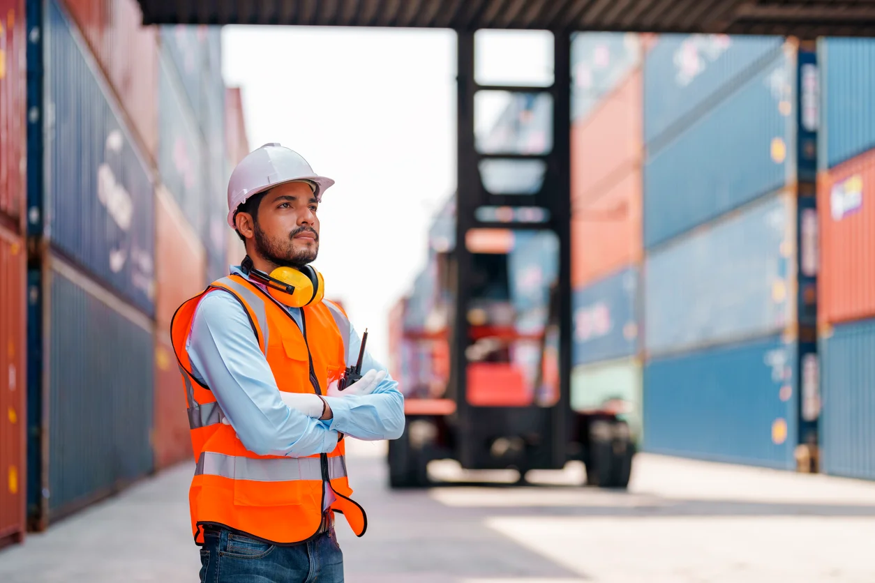 Trident Law HQ represents dockworkers injured on duty, as represented by a man in a hard hat and orange vest standing next to a piece of heavy machinery in a shipping yard 