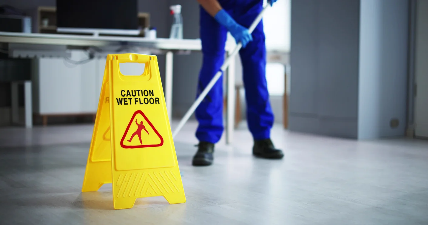 A close-up shot of a yellow wet floor sign and a worker in blue mopping up in the background representing slip and fall injury cases pursued by Trident Law HQ